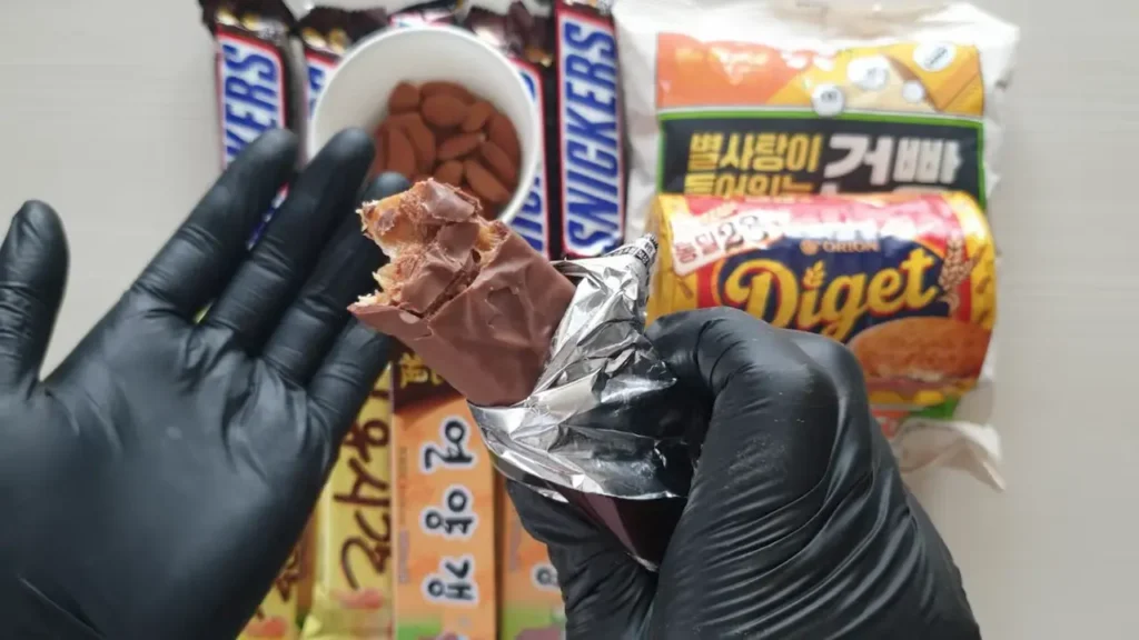 흑색 장갑을 낀 손으로 한 입 베어 문 스니커즈 초콜릿 바를 들고 있는 모습. 배경에는 양갱, 다이제, 아몬드 등 재난 시 즉각 섭취 가능한 다양한 행동식(Action Food) 비상식들이 배치되어 있음