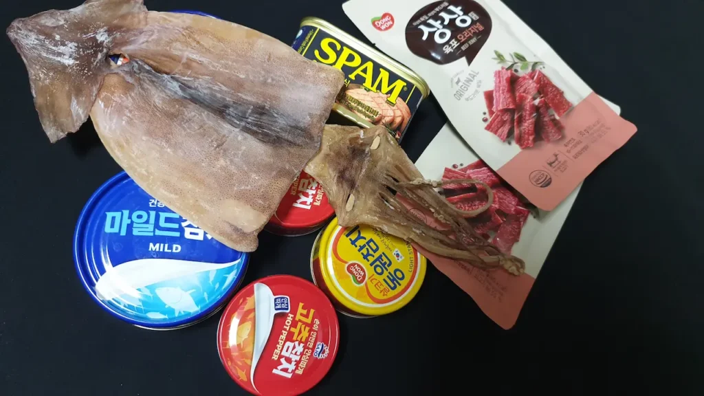 말린 오징어, 소고기 육포 파우치, 그리고 참치와 스팸 통조림들이 모여 있는 모습으로 장기 재난 시 필요한 동식물성 단백질 비상식량 구성을 보여주는 이미지